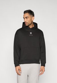 Sweatshirt preta com capuz, bolso frontal tipo canguru, com um pequeno logotipo branco no peito e punhos canelados. modelo calças de fato de treino cinzentas.