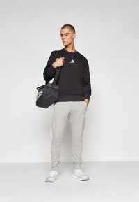 Sort sweatshirt med hvid Adidas-logo, grå sweatpants med hvide striber og hvide sneakers. Model holder en sort Adidas taske.