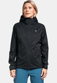 Schwarze wasserdichte Jacke mit einer passgenauen Kapuze, Frontreißverschluss und grauen Logos, kombiniert mit blauen Hosen, die eine glatte matte Oberfläche aufweisen.