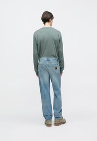 Zöld hosszú ujjú ing és bővebb szabású világoskék farmernadrág, barna bőrfolttal a hátsó zseben. A modell olíva zöld csizmát visel.