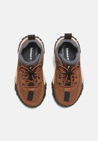 Zapatos marrones de múltiples materiales con un upper texturizado, suelas de goma negra, perforaciones y cordones elásticos para un ajuste seguro. Marca Timberland visible.