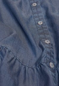 Himmelblaue Chambray-Bluse mit einer Knopfleiste aus Perlenknöpfen, versammelten Details am Saum und einem glatten, leicht strukturierten Stoff.