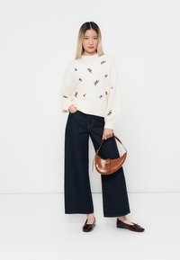 Frau trägt einen weißen, blumigen Pullover, weite, dunkle Jeans, burgunderfarbene Ballerinas und hält eine braune, strukturierte Handtasche vor einem weißen Hintergrund.
