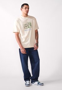 T-shirt crème à manches courtes avec un graphique de montagne verte et du texte bleu, associé à un jean en denim bleu décontracté et des baskets marines.