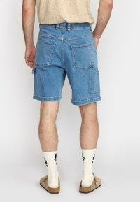 Lichte blauwe denim short met een ontspannen pasvorm, omgeslagen zoom en twee achterzakken met stikseldetails. Draagt samen met witte sokken en instappers.