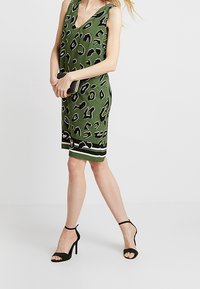 Vestido sin mangas verde con estampado de leopardo negro, diseño con escote en V, con un dobladillo sólido de rayas blancas y negras, combinado con sandalias de tacón negras.