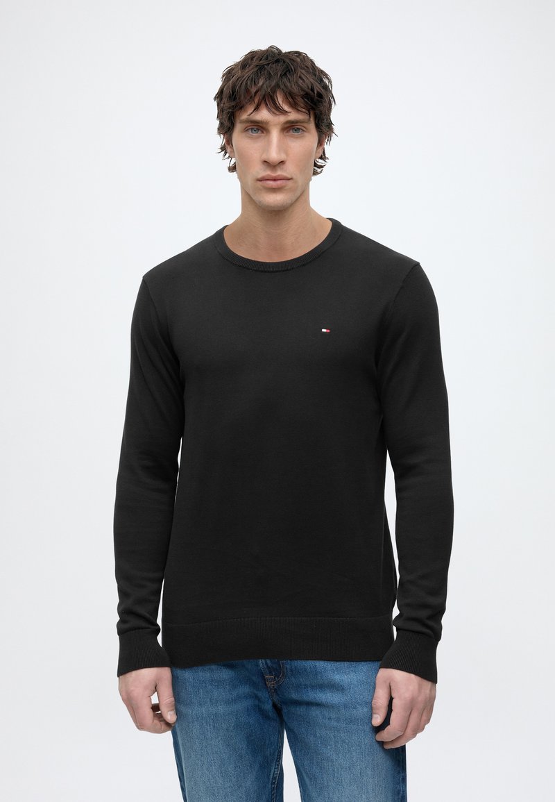 Homme aux cheveux bouclés portant un pull noir à manches longues col rond et un jean bleu, face à un fond clair et uni.