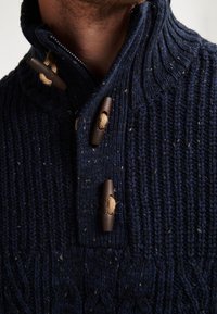 Pulover tricotat de culoare bleumarin, cu guler înalt, având nasturi toggle din lemn închis la culoare și un model texturat cu striații pe întreaga suprafață.