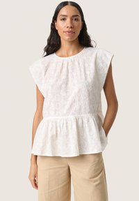 Vit peplumtop med blommig brodyr, korta fjärilsärmar och en gathered neckline; kombinerad med ljusbeiga byxor.