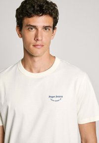 Balta medvilnės marškinėliai su trumpomis rankovėmis ir apvalia iškirpte. Išskirtinė mėlyna siuvinėta "Pepe Jeans" emblema ant kairiojo krūtinės.