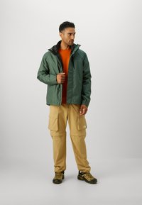 Chaqueta impermeable verde con interior negro, camisa naranja, pantalones cargo beige con múltiples bolsillos y zapatos de senderismo grises.