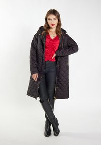 Cappotto lungo trapuntato nero con cappuccio, dettagli dorati, abbinato a una blusa rossa arricciata e pantaloni neri, indossato con stivali neri al ginocchio.