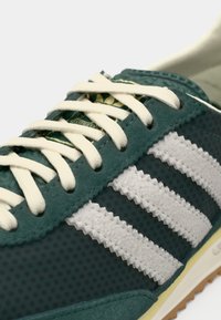 Grøn sneaker med hvide snørebånd og tre hvide stribede detaljer på siden, tæt billede af den øverste del og tungen.