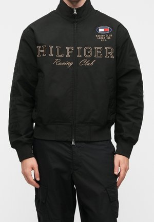 Personne portant une veste noire à fermeture éclair complète avec le texte "HILFIGER Racing Club" et un petit écusson du club de course sur la poitrine, associée à un pantalon cargo noir.