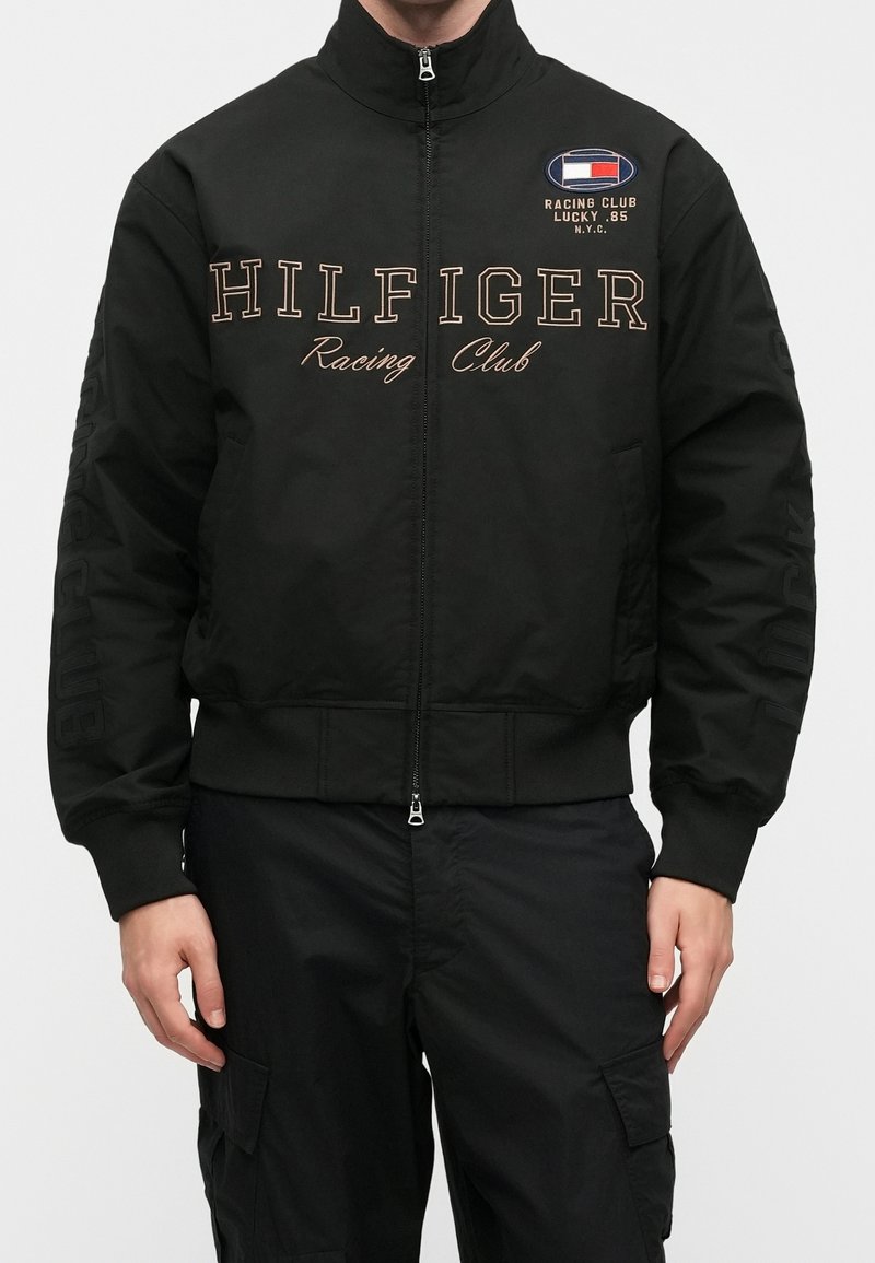 Personne portant une veste noire à fermeture éclair complète avec le texte "HILFIGER Racing Club" et un petit écusson du club de course sur la poitrine, associée à un pantalon cargo noir.