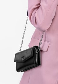 Schwarze Leder-Crossbody-Tasche mit einem Klappendesign, silberner Kettenriemen und Reißverschlussfach, präsentiert vor einem pinken Kleidungsstück.