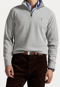 Sweat-shirt gris à fermeture éclair avec manches longues, arborant un logo discret sur la poitrine, accompagné d'un pantalon en velours côtelé marron et d'une ceinture marron.