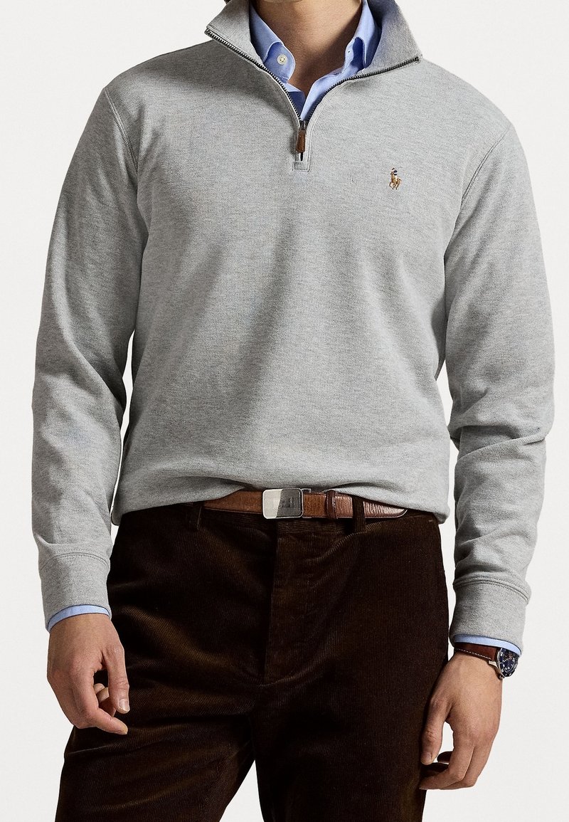 Sweat-shirt gris à fermeture éclair avec manches longues, arborant un logo discret sur la poitrine, accompagné d'un pantalon en velours côtelé marron et d'une ceinture marron.