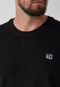 Bearded man wearing a black sweatshirt with ribbed collar and a small "s.O" logo patch on the chest.