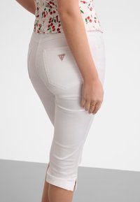 Femme portant un pantalon blanc court avec des ourlets retroussés et un haut sans manches à motif cerises, debout avec un bras détendu le long du corps.