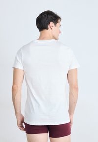 Homme portant un t-shirt blanc uni à manches courtes et un short bordeaux, se tenant de dos contre un fond clair.