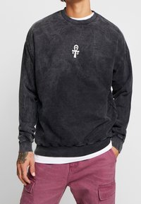 Sudadera gris oscuro en tela suave y texturizada. Presenta un símbolo ankh blanco en el pecho y puños y cintura acanalados.