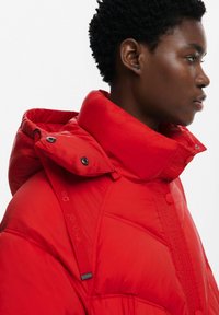 Chaqueta de plumas roja con un alto cuello, acentos de tejido texturizado y botones negros. Presenta una correa de marca en el hombro y una costura lateral.