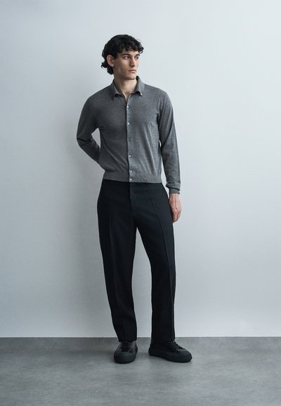 Homme debout contre un mur uni portant un cardigan gris boutonné, un pantalon noir et des baskets noires, regardant sur le côté avec une main derrière le dos.