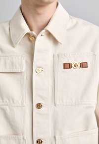 Beige bomuldsjakke med guldknapper, to frontlommer og en dekorativ brun læderstrop med en rund gul accent.