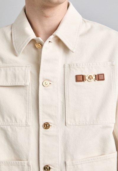 Beige cotton jacket featuring gold buttons, two front pockets, and a decorative brown leather strap with a round gold accent.