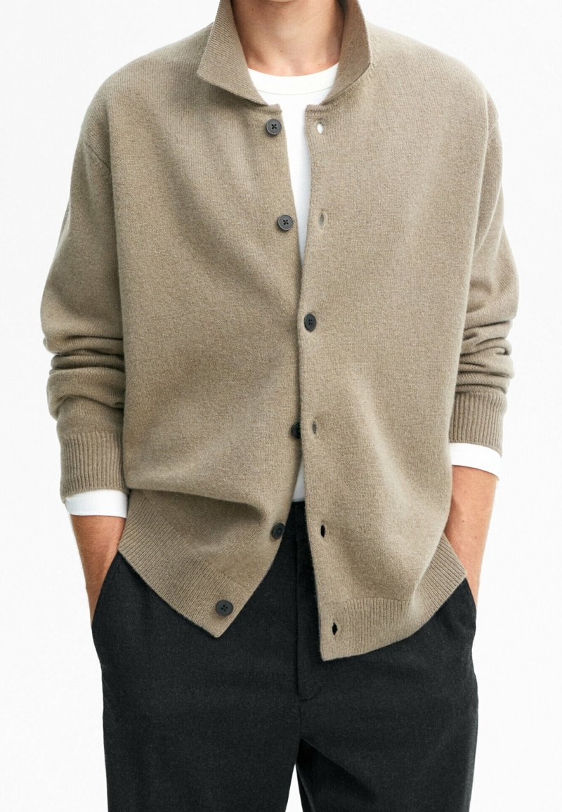 Homme portant un cardigan beige boutonné par-dessus une chemise blanche à manches longues, les mains dans les poches, et un pantalon noir, sur fond blanc.