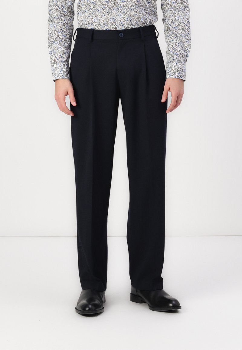 Pantaloni eleganți bleumarin, cu pliuri, dintr-un material neted. Au curea de prindere și închidere cu nasture, asortați cu pantofi negri.