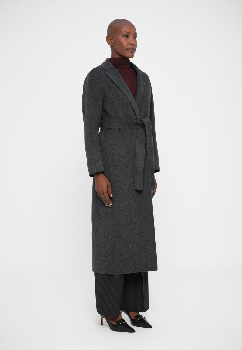 Woman standing wearing a long dark grey belted coat, burgundy turtleneck, pinstriped wide-leg trousers, and black pointed heels.