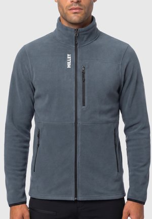 Mann trägt eine blaue Fleecejacke mit durchgehendem Reißverschluss, Reißverschlusstaschen an Brust und Seiten sowie dem Markennamen "Millet" auf der linken Brust.