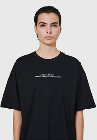 Person med kort mörkt hår som bär en svart oversize t-shirt med vit text "OFF DUTY GEAR | HALO 1968|972|981" på bröstet, neutral bakgrund.