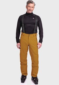 Ski pants in mustard yellow, made of durable material. Features adjustable black suspenders, zip pockets, and a sleek, functional design.