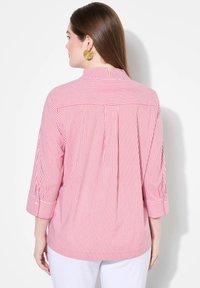 Maglietta a righe rosse e bianche con colletto, maniche a tre quarti e piega sul retro. Il tessuto appare liscio e leggero.