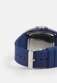 Bracelet de montre en silicone bleu avec une finition mate, doté d'une boucle en acier inoxydable argenté et d'un boîtier rectangulaire avec du matériel visible.