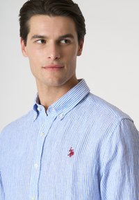 Camicia a righe blu e bianche con bottoni, dotata di un piccolo logo rosso ricamato sul petto, caratterizzata da un colletto classico e un tessuto strutturato.