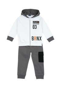 Veste à capuche zippée blanche avec des poignets gris, ornée de texte noir et orange. Pantalon de survêtement gris avec une poche cargo noire. Matière en coton.