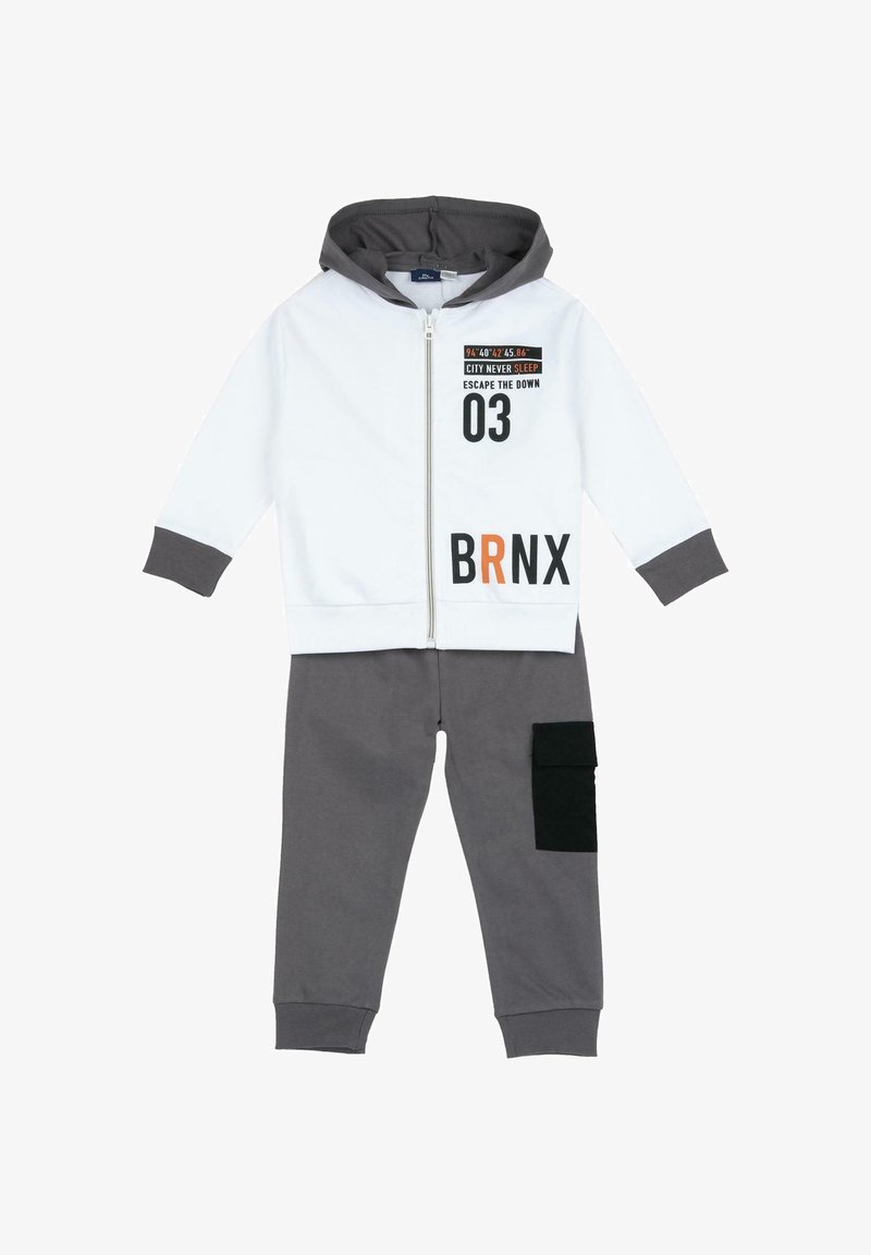 Veste à capuche zippée blanche avec des poignets gris, ornée de texte noir et orange. Pantalon de survêtement gris avec une poche cargo noire. Matière en coton.