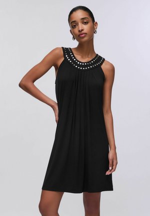 Donna con capelli scuri raccolti indossa un abito nero senza maniche con borchie argentate a forma circolare lungo la scollatura, posando con una mano sul fianco.