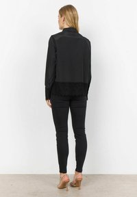 Black collared blouse with soft fabric and lace trim at the hem, paired with black fitted trousers and open-toe sandals.