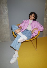 Veste bomber rose, haut en tricot blanc, jean flare bleu clair et baskets blanches avec des accents roses, assis dans un environnement jaune et gris.