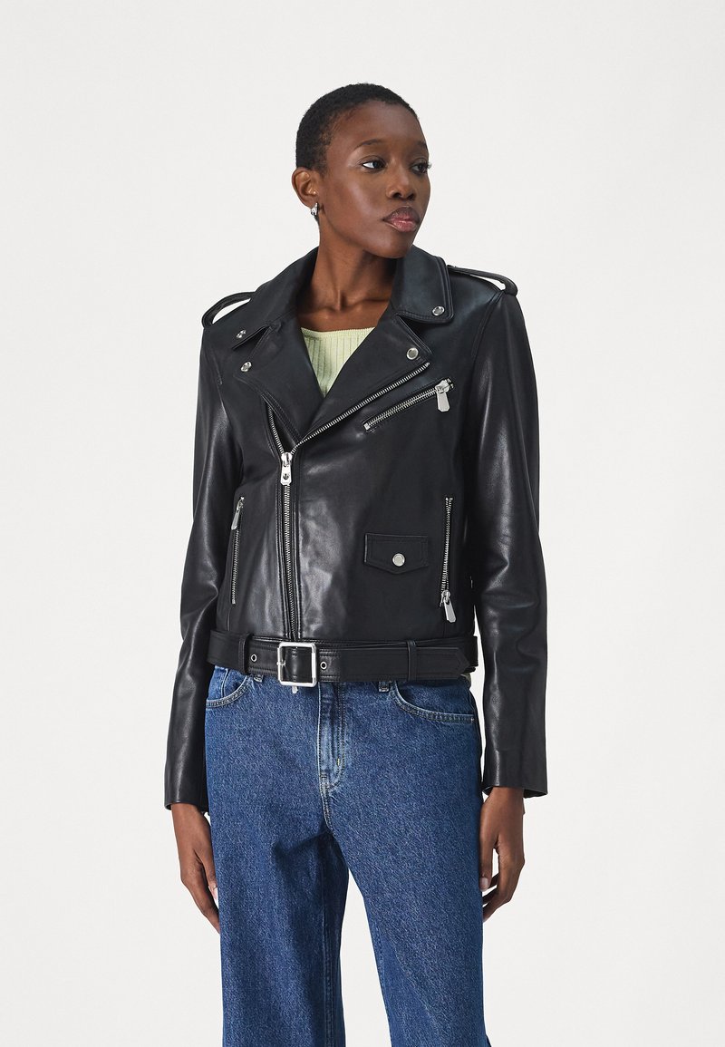 Crna kožna moto jakna s asimetričnim patentnim zatvaračem, epauletama i prednjim džepovima, kombinirana sa zelenom majicom i visokim plavim trapericama.