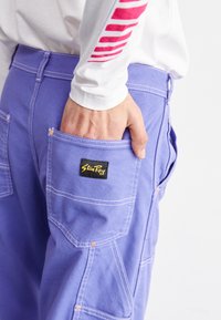 Purple work pants feature a patch pocket with a black label, orange stitching details, and a straight fit. A white long-sleeve shirt is partially visible.