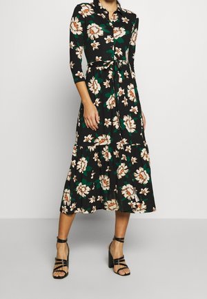 Schwarzes Blumenkleid mit beigen und grünen Blumen, einem Kragen, dreiviertel Ärmeln und einem Stufenrock. Kombiniert mit schwarzen High-Heel-Sandalen.