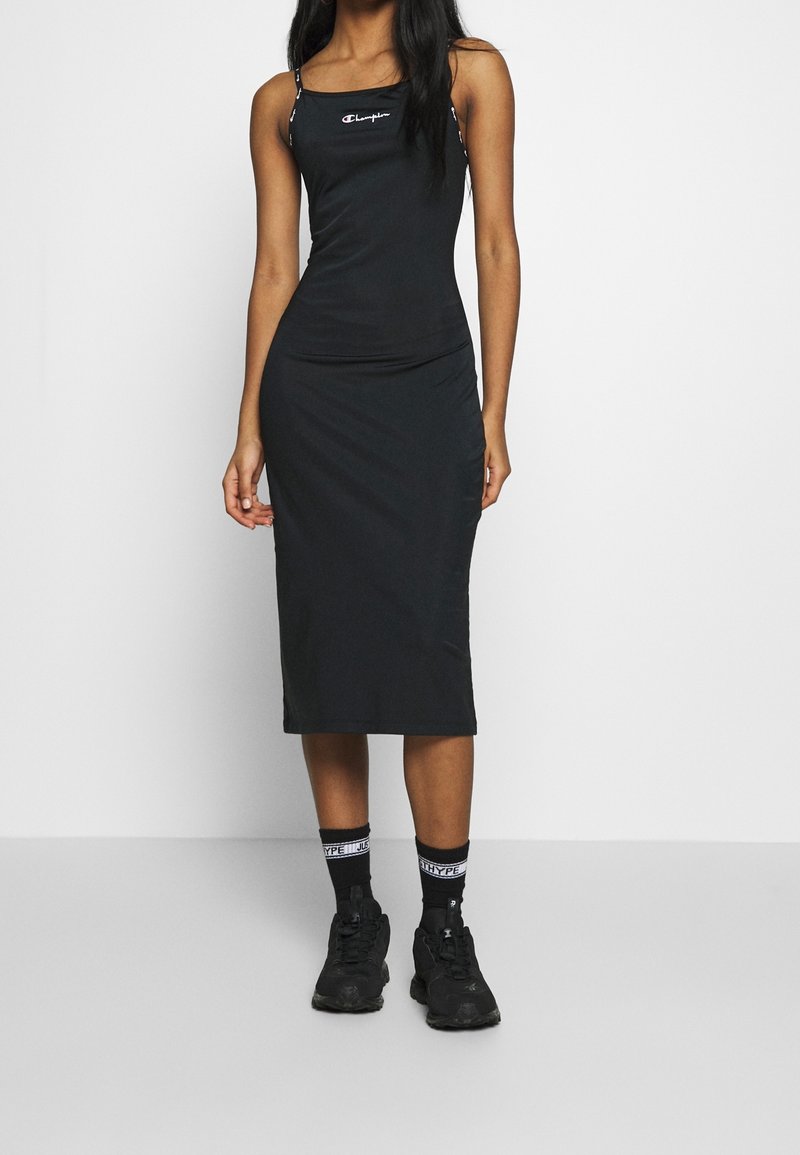 Femme portant une robe midi noire ajustée avec de fines bretelles, des baskets noires et des chaussettes noires avec un texte blanc, debout devant un fond blanc.