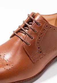 Chaussure en cuir marron avec une finition lisse, des accents perforés et un design classique, dotée de lacets marron et d'une forme de bout arrondi.