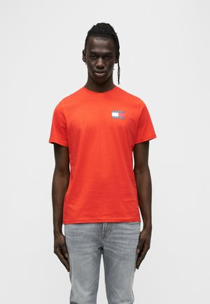 Camiseta de algodón roja con cuello redondo y mangas cortas. Presenta un parche con logo azul y blanco en el pecho izquierdo. Combinada con jeans grises.